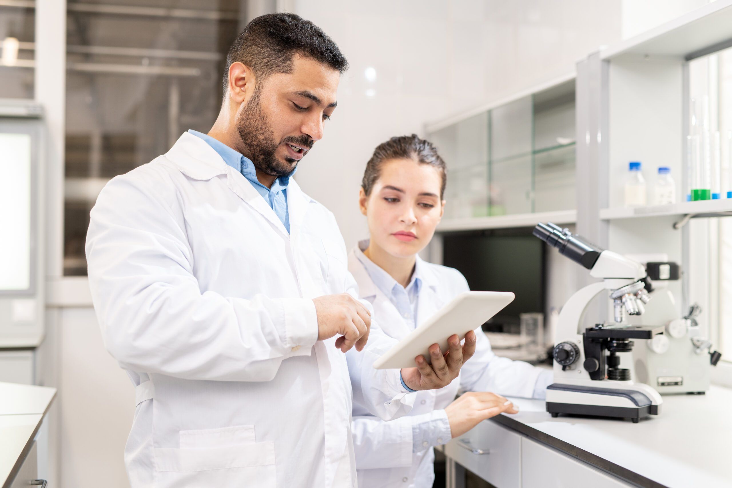 Confident experienced middle-eastern scientist in lab coat showing data on tablet to colleague while they examining test solution in laboratory