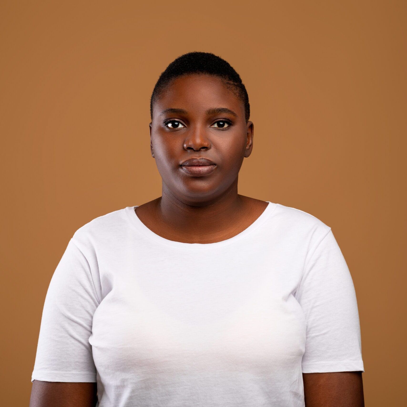 Portrait Of Serious African American Young Woman Wearing White T-Shirt Posing Looking At Camera Standing Isolated Over Brown Studio Background. Female Headshot Of Black Lady